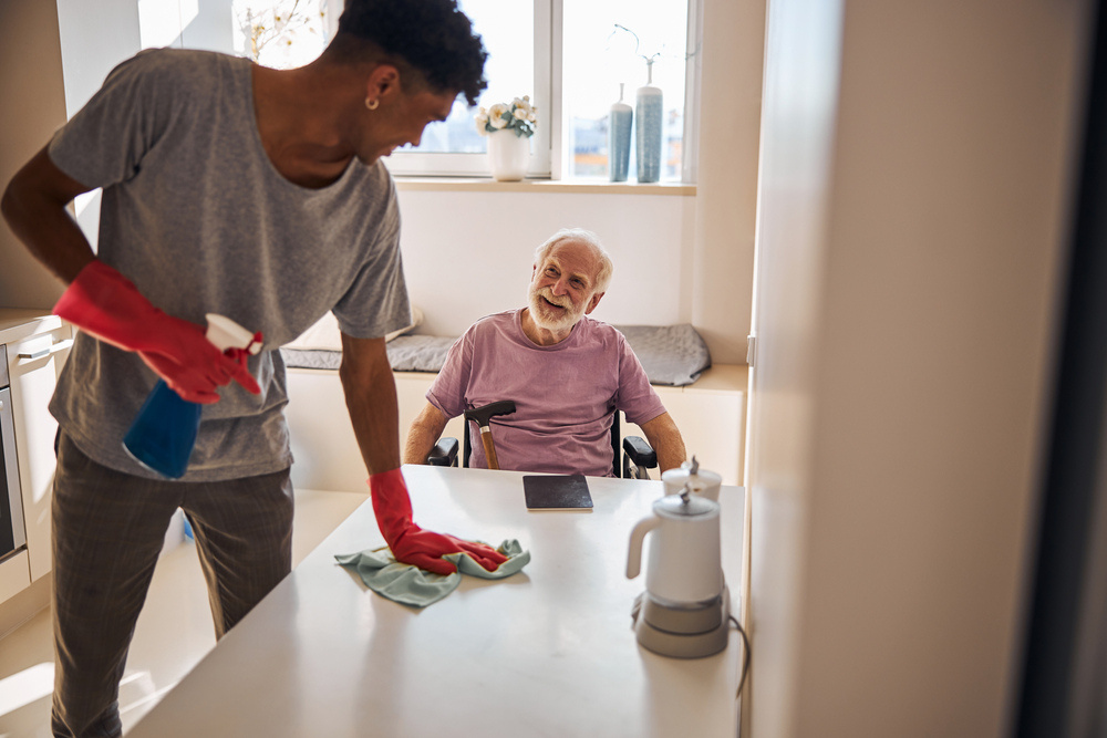 A friendly companion wiping down the kitchen table while chatting with a client