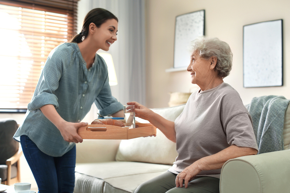 A companion bringing breakfast on a tray to a smiling older lady at home